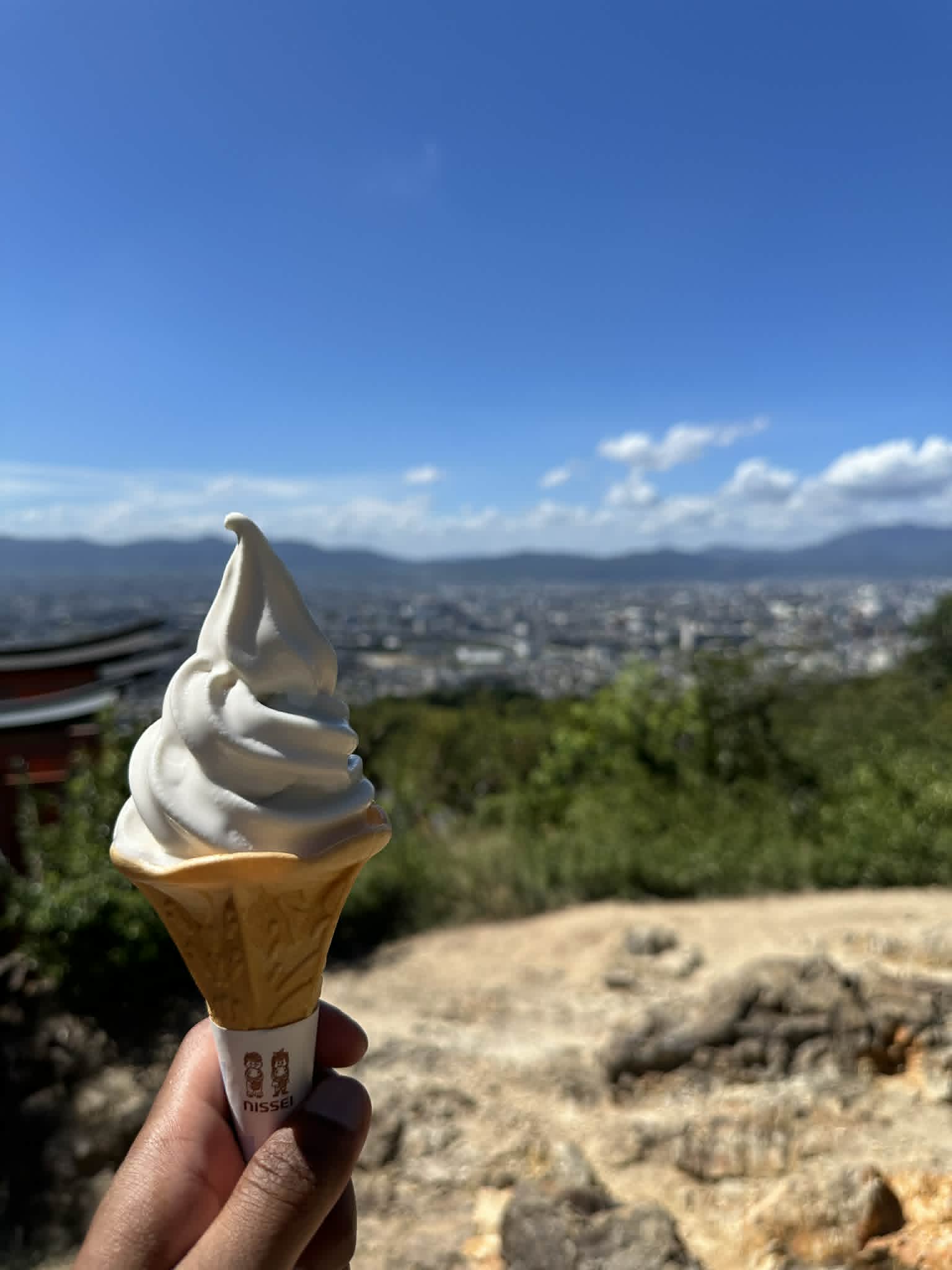 A vanilla ramune-flavour soft serve cone held up against a panoramic view of Kyoto from Fushimi Inari shrine in September
