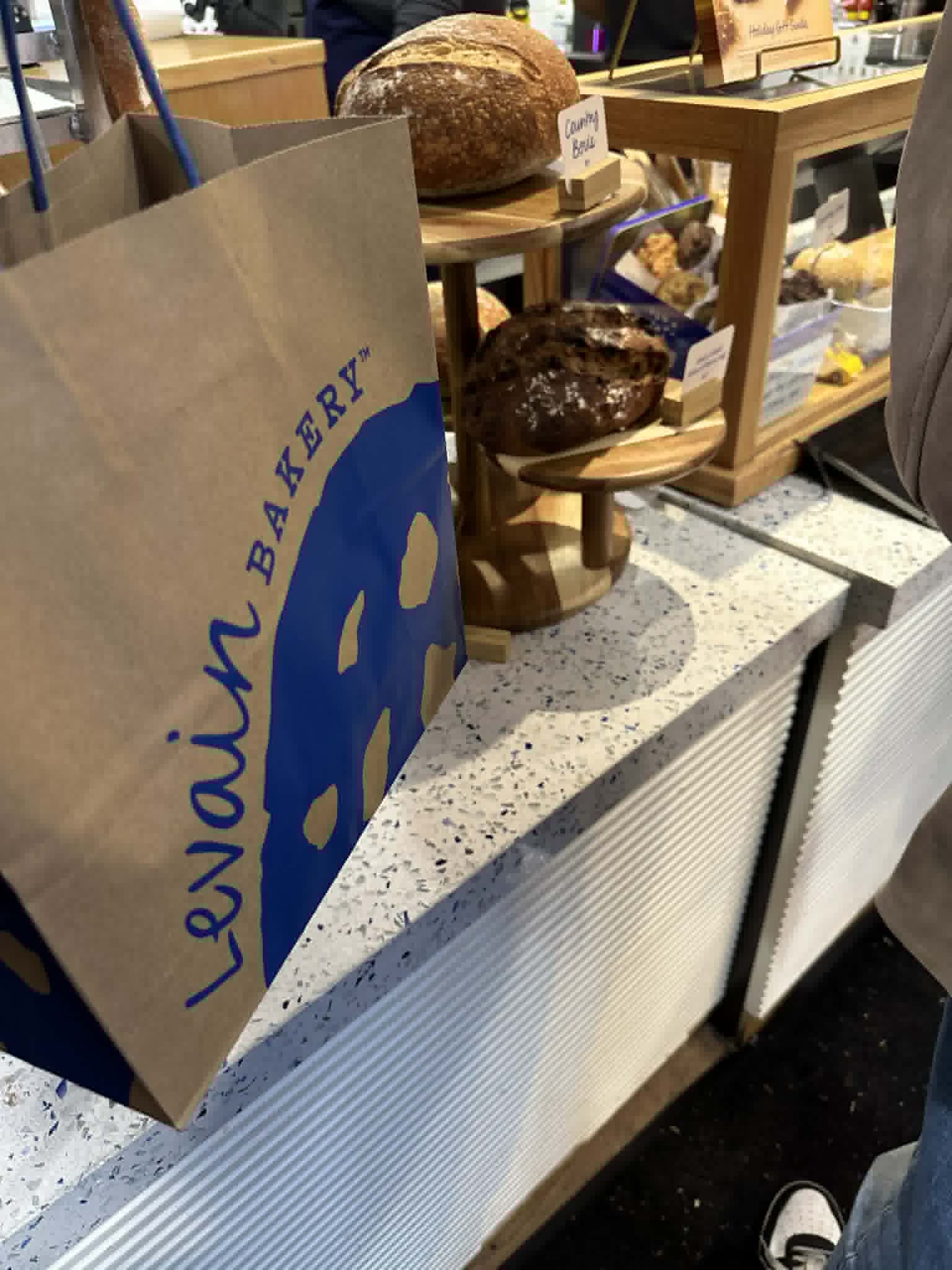 Levain Bakery paper bag in foreground with rustic bread loaves displayed on the bakery counter behind, NYC