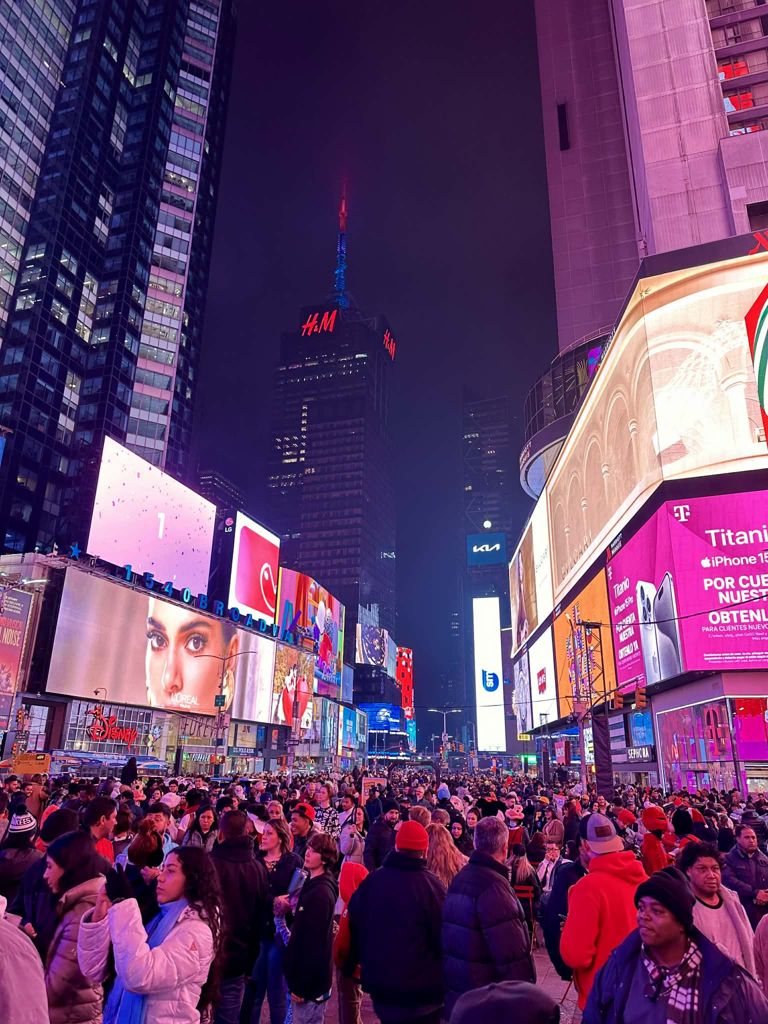 Times Square at night in late December with crowds of people, illuminated billboards on every building, and the H&M sign on the central tower