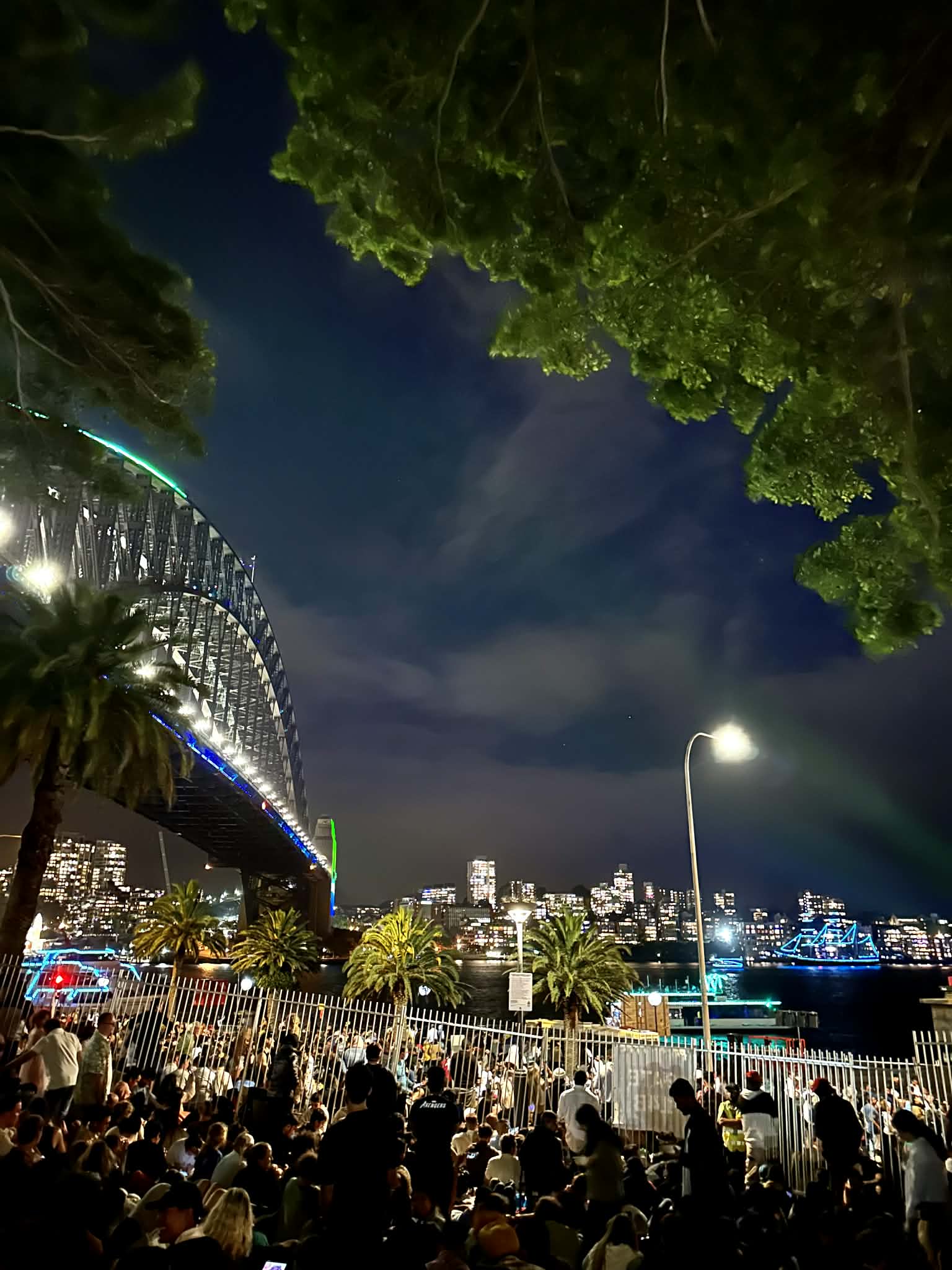 Sydney Harbour Bridge illuminated in green and blue lights at night, with a large crowd waiting for New Year's Eve fireworks