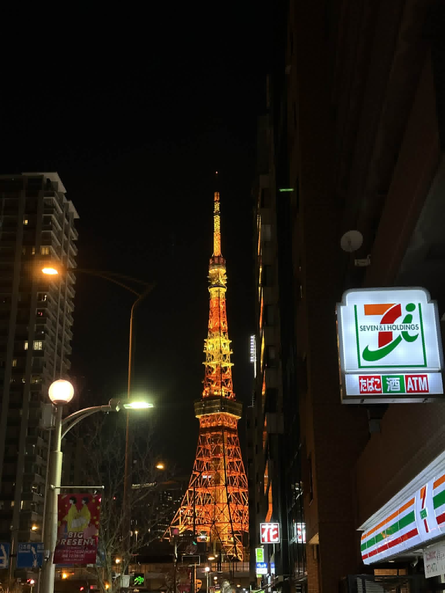 Tokyo Tower lit up orange at night, framed between buildings with a 7-Eleven konbini sign in the foreground